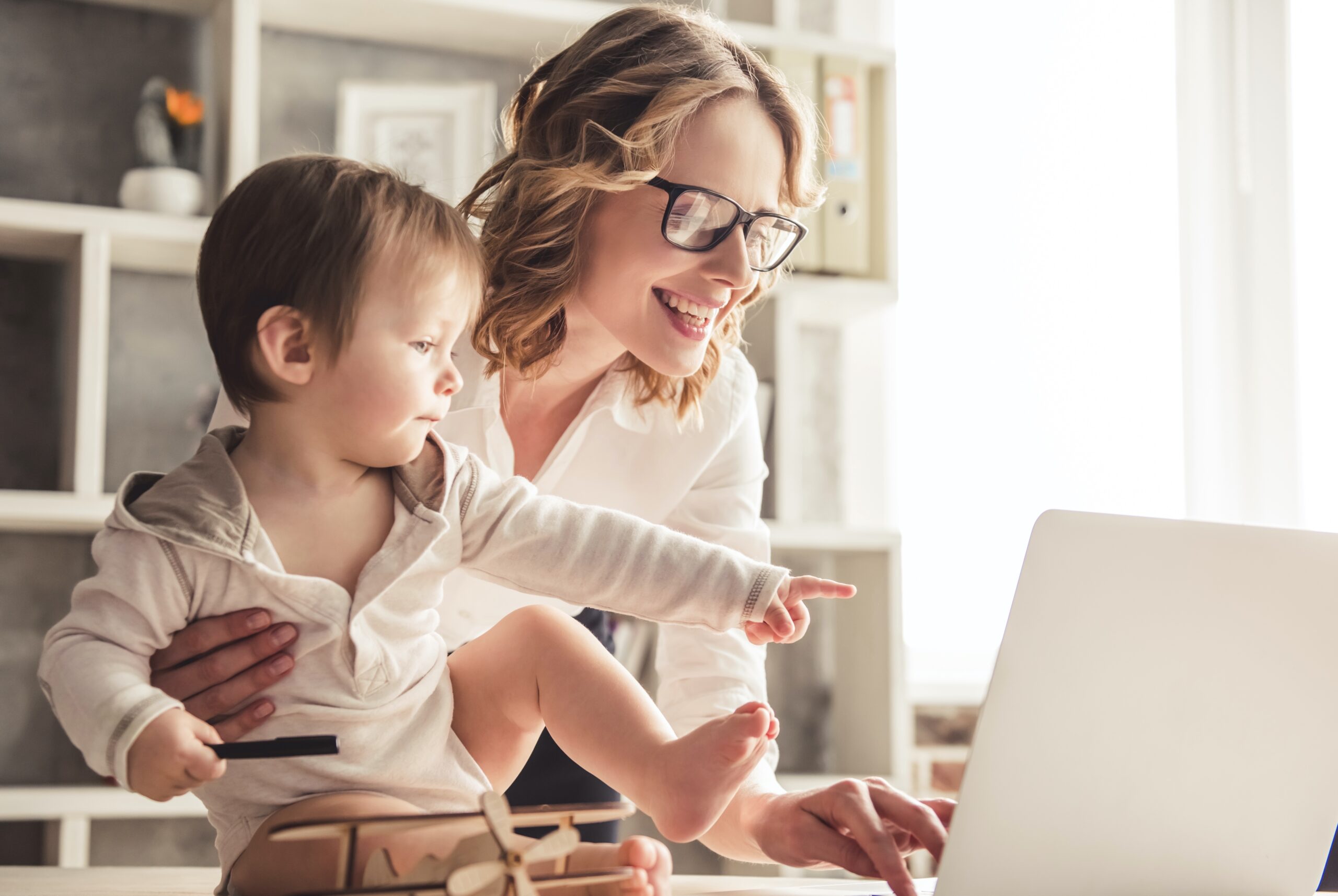 Beautiful business mom is using a laptop and smiling while spending time with her cute baby boy at home